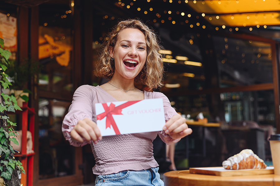 Young Happy Smiling Woman In Casual Clothes Sitting Alone At Table In Coffee Shop Cafe Eat Breakfast Hold Gift Certificate Coupon Voucher Card